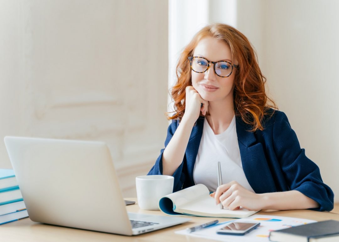 A thoughtful woman with curly red hair sits at a desk, wearing glasses, surrounded by books and a laptop, writing in a notebook.